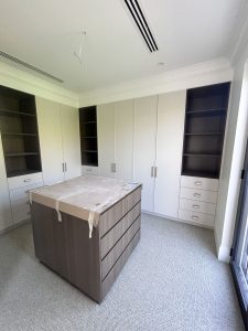 Luxury walk-in wardrobe in Rose Bay featuring built-in cabinetry, open shelving, drawer storage, and a central timber island with stone benchtop.